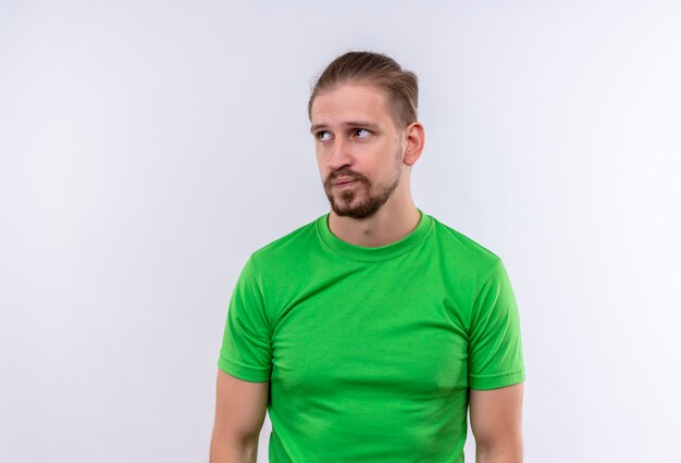 Joven guapo en camiseta verde mirando a un lado disgustado y confundido de pie sobre fondo blanco.