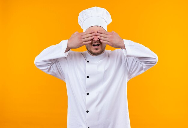 Joven guapo cocinero en uniforme de chef cerrando los ojos con las manos con la boca abierta aislada en el espacio naranja