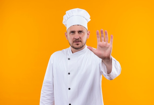 Joven guapo cocinero en uniforme de chef haciendo un gesto de parada y mirando aislado en el espacio naranja