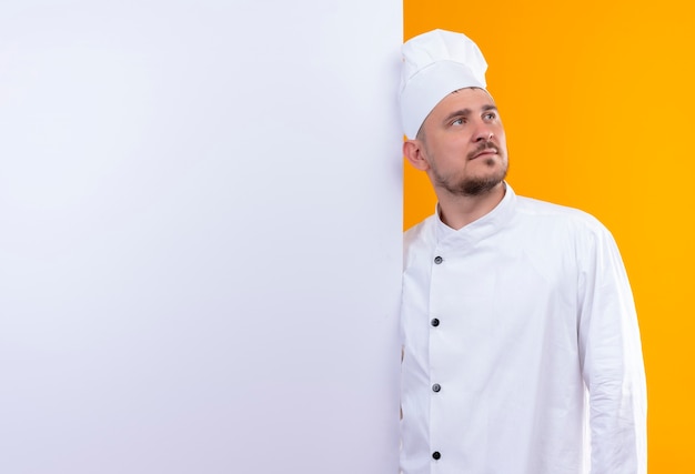 Joven guapo cocinero en uniforme de chef de pie detrás de una pared blanca mirando directamente aislado en el espacio naranja