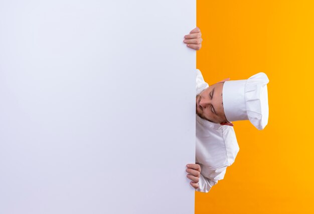 Joven guapo cocinero en uniforme de chef de pie detrás de una pared blanca y mirándolo aislado en el espacio naranja