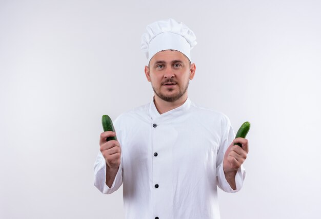 Joven guapo cocinero en uniforme de chef sosteniendo pepinos mirando aislado en el espacio en blanco