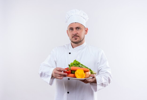 Joven guapo cocinero en uniforme de chef sosteniendo un plato con verduras en un espacio en blanco aislado