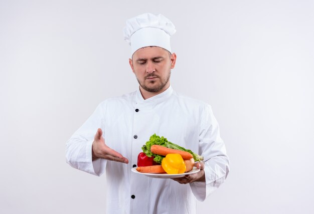 Joven guapo cocinero en uniforme de chef sosteniendo un plato con verduras mirando y señalando en un espacio en blanco aislado