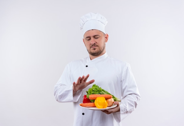 Joven guapo cocinero en uniforme de chef sosteniendo un plato con verduras mirándolos y manteniendo la mano sobre ellos en un espacio en blanco aislado