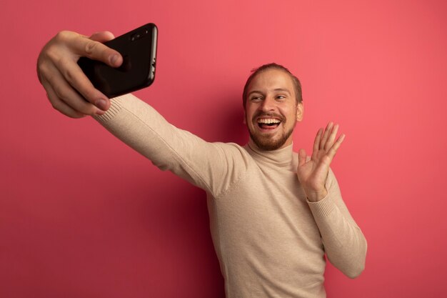 Foto gratuita joven guapo en cuello alto beige con smartphone haciendo selfie feliz y positivo saludando con la mano