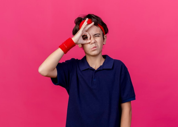 Joven guapo deportivo con diadema y muñequeras con aparatos dentales guiñando un ojo en la parte delantera y haciendo gesto de mirada aislado en la pared rosa