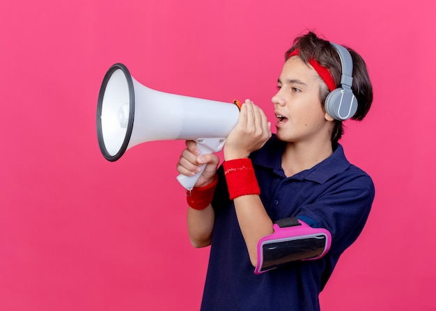 Joven guapo deportivo con diadema y muñequeras y auriculares brazalete de teléfono con aparatos dentales mirando directamente hablando por altavoz aislado en pared carmesí