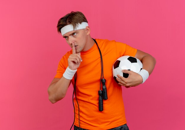 Joven guapo deportivo con diadema y muñequeras con saltar la cuerda alrededor del cuello sosteniendo un balón de fútbol mirando a un lado y gesticulando silencio aislado sobre fondo rosa con espacio de copia