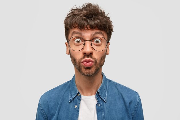 Un joven guapo y emotivo hace pucheros con asombro, mira fijamente a través de grandes gafas, tiene una barba oscura, vestido con una camisa de mezclilla, posa contra la pared blanca