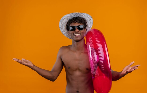 Un joven guapo enojado de piel oscura con cabello rizado con sombrero para el sol sosteniendo un anillo de piscina inflable