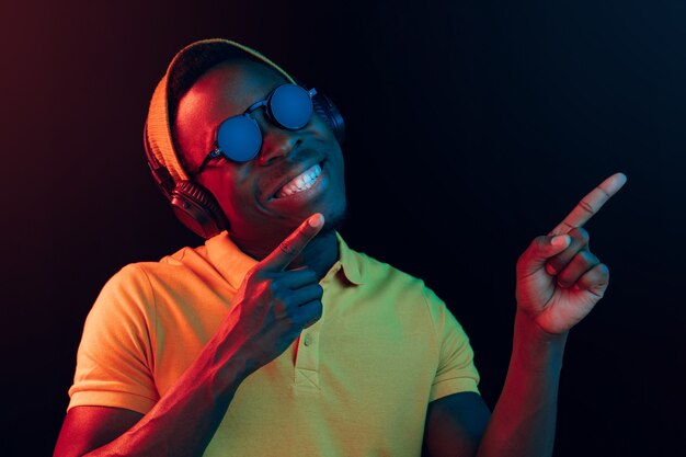 El joven guapo feliz hipster hombre escuchando música con auriculares en estudio negro con luces de neón. Discoteca, club nocturno, estilo hip hop, emociones positivas, expresión facial, concepto de baile