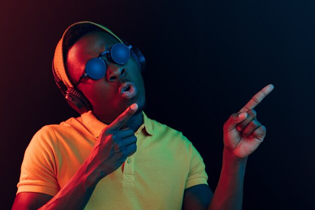 El joven guapo feliz hipster hombre escuchando música con auriculares en estudio negro con luces de neón.