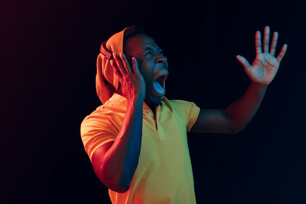 El joven guapo feliz sorprendido hipster hombre escuchando música con auriculares en el estudio negro con luces de neón. Discoteca, club nocturno, estilo hip hop, emociones positivas, expresión facial, concepto de baile