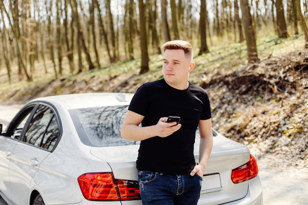 Joven guapo, feliz, usa el teléfono inteligente en el automóvil y al aire libre