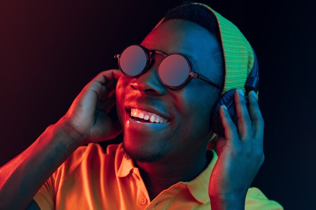 El joven guapo hipster feliz escuchando música con auriculares en el estudio negro con luces de neón. Discoteca, club nocturno, estilo hip hop, emociones positivas, expresión facial, concepto de baile
