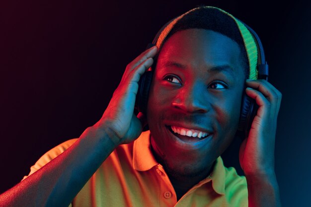 El joven guapo hipster feliz escuchando música con auriculares en el estudio negro con luces de neón