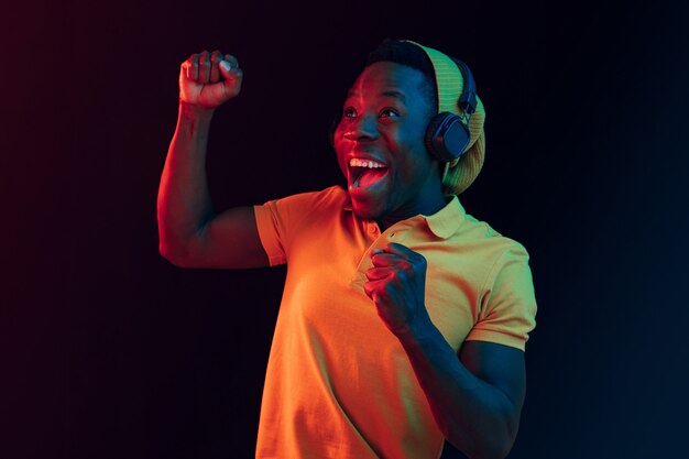 El joven guapo hipster feliz escuchando música con auriculares en el estudio negro con luces de neón