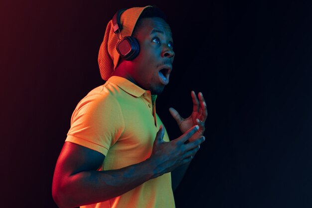 El joven guapo hipster feliz escuchando música con auriculares en el estudio negro con luces de neón