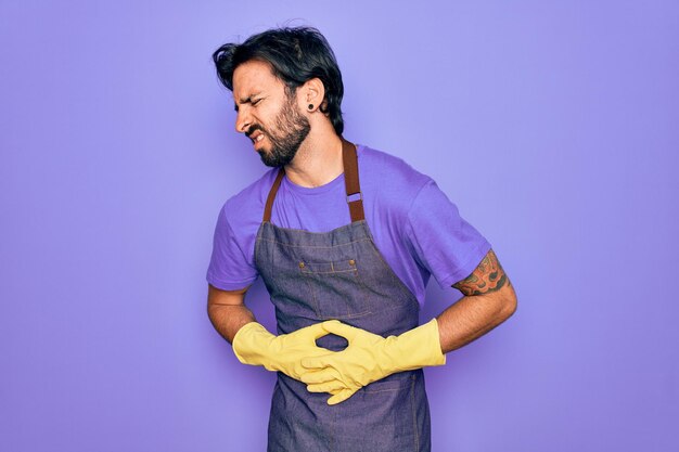 Joven guapo hispanic clenaer hombre vestido con delantal de tareas domésticas y guantes de lavado con la mano en el estómago porque náuseas enfermedad dolorosa sensación de malestar concepto de Ache