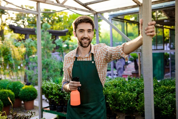 Joven guapo jardinero alegre sonriendo, posando entre plantas y flores