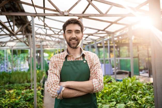 Joven guapo jardinero sonriendo, posando con los brazos cruzados entre flores