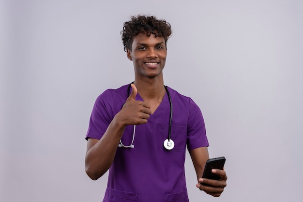 Un joven y guapo médico de piel oscura con cabello rizado y uniforme violeta con estetoscopio sosteniendo su teléfono inteligente mientras muestra un gesto de ok