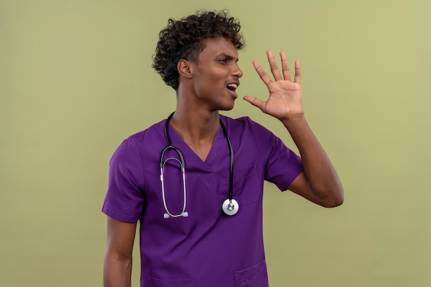 Un joven y guapo médico de piel oscura enojado con cabello rizado vistiendo uniforme violeta con estetoscopio llamando a alguien mientras mira de lado en un espacio verde