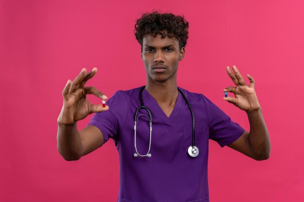 Un joven y guapo médico de piel oscura enojado con cabello rizado vistiendo uniforme violeta con estetoscopio sosteniendo pastillas