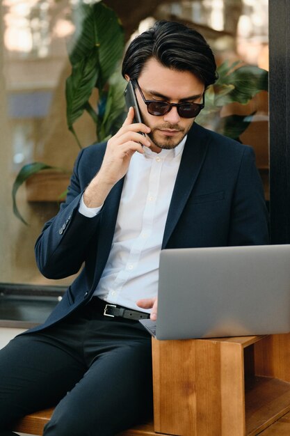 Joven guapo moreno barbudo con camisa blanca, chaqueta clásica y gafas de sol hablando por teléfono celular trabajando cuidadosamente en una computadora portátil mientras está sentado en un café en la calle