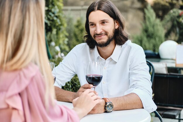 Joven guapo moreno barbudo en camisa con una copa de vino mirando felizmente a su novia en una cita romántica en un café al aire libre