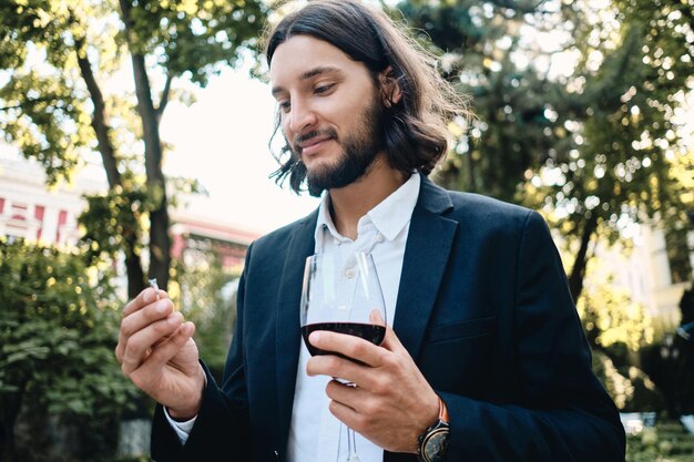 Joven guapo moreno barbudo con una copa de vino sosteniendo soñadoramente el anillo de bodas en el restaurante al aire libre