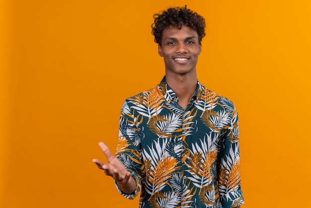 Foto gratuita un joven guapo de piel oscura con cabello rizado en hojas de camisa estampada sonriendo y levantando la mano