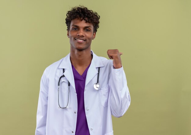 Un joven guapo de piel oscura con pelo rizado vestido con bata blanca con estetoscopio sonriendo apuntando con el dedo pulgar sobre un espacio verde