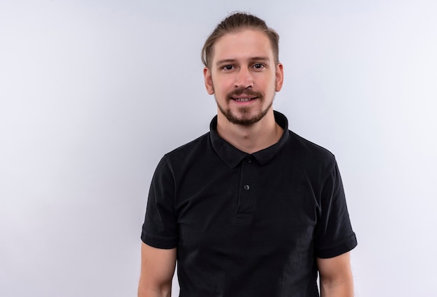 Foto gratuita joven guapo en polo negro sonriendo amable mirando a la cámara de pie sobre fondo blanco.