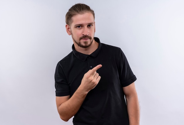 Joven guapo en polo negro sonriendo confiado apuntando hacia el lado con el dedo parado sobre fondo blanco.