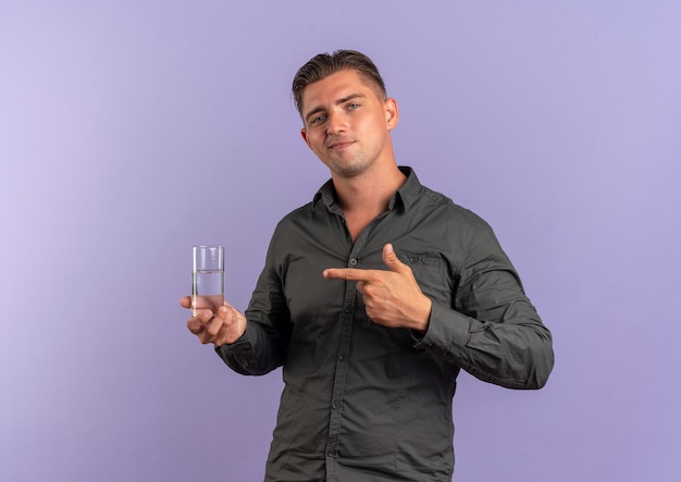 Foto gratuita joven guapo rubio contento sostiene y apunta a un vaso de agua aislado en el espacio violeta con espacio de copia