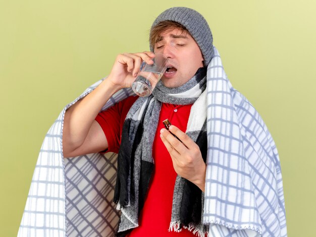 Joven guapo rubio enfermo con gorro de invierno y bufanda envuelto en cuadros con medicamento en vaso vaso de agua aislado en la pared verde oliva