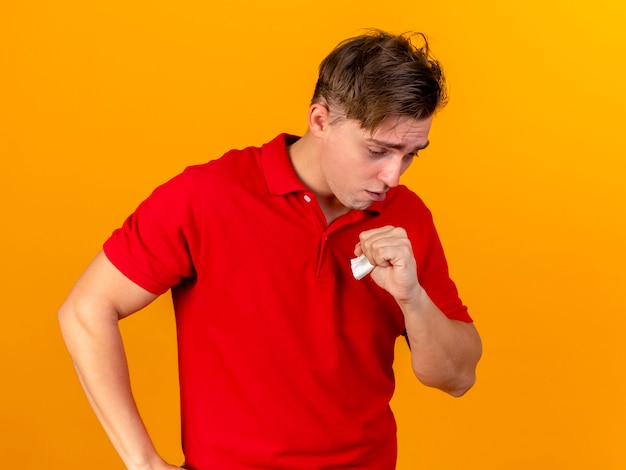 Joven guapo rubio enfermo manteniendo la mano en la cintura mirando hacia abajo tosiendo aislado en la pared naranja con espacio de copia