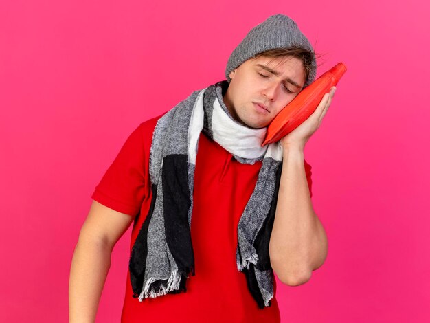 Joven guapo rubio enfermo con sombrero de invierno y bufanda sosteniendo una botella de agua caliente tocando la cara con los ojos cerrados aislados en la pared rosa