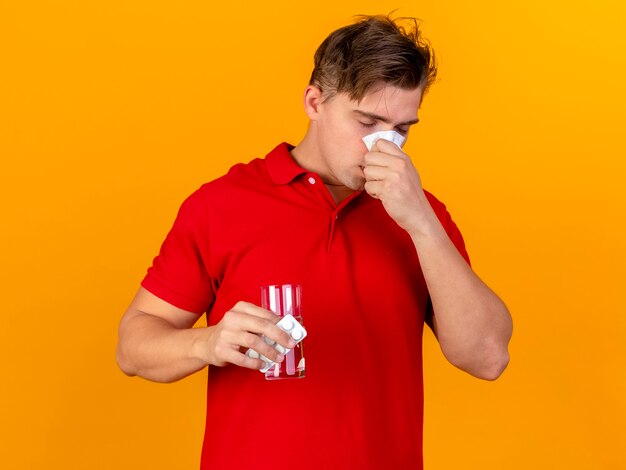 Joven guapo rubio enfermo sosteniendo un paquete de tabletas médicas y un vaso de agua limpiando la nariz con una servilleta con los ojos cerrados aislados sobre fondo naranja