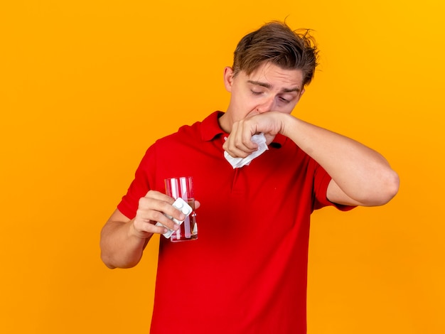 Foto gratuita joven guapo rubio enfermo sosteniendo un paquete de tabletas médicas y un vaso de agua con una servilleta limpiando la nariz aislada en la pared naranja