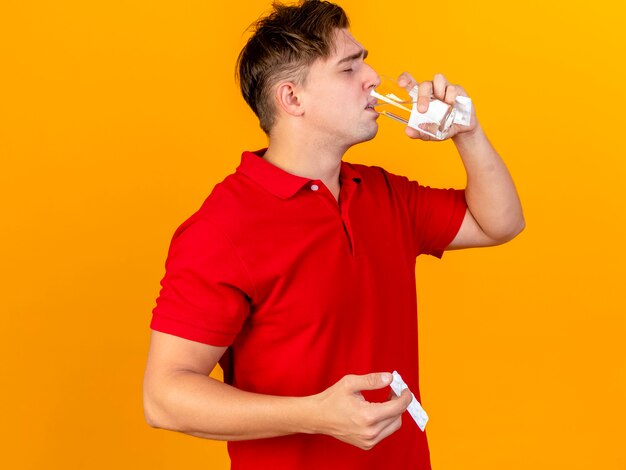 Joven guapo rubio enfermo sosteniendo una servilleta tomando la píldora bebiendo agua de vidrio aislado en la pared naranja