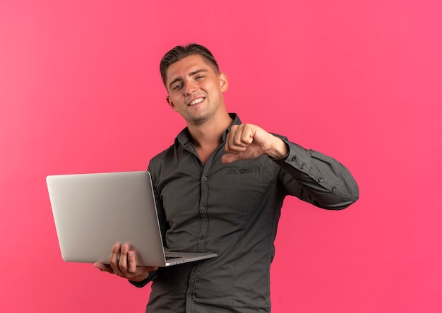 Joven guapo rubio seguro sostiene portátil y apunta a sí mismo aislado sobre fondo rosa con espacio de copia
