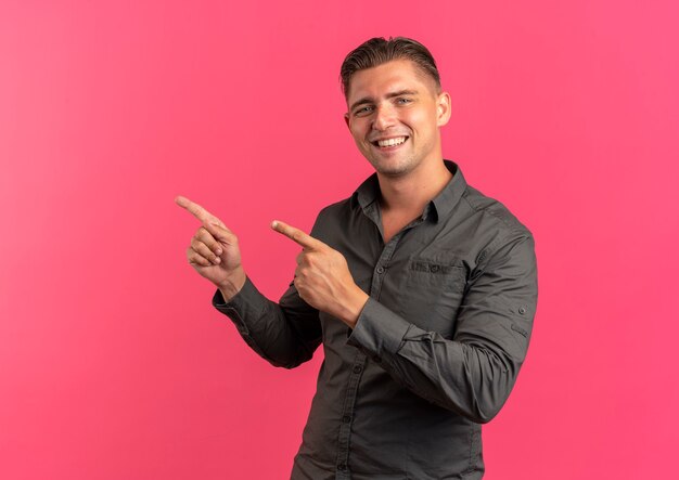 Joven guapo rubio sonriente apunta al lado mirando a cámara aislada sobre fondo rosa con espacio de copia