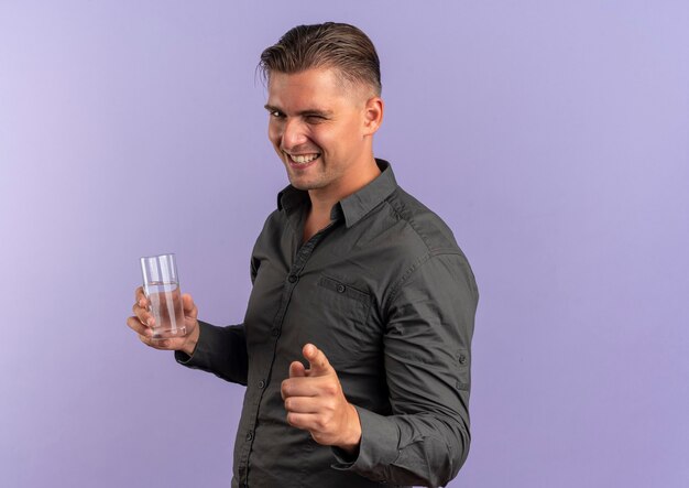 Joven guapo rubio sonriente parpadea y apunta a la cámara sosteniendo un vaso de agua aislado sobre fondo violeta con espacio de copia