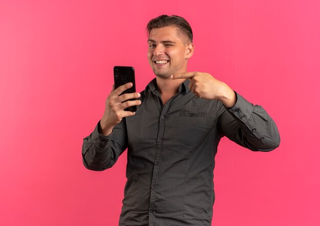 Joven guapo rubio sonriente parpadea sosteniendo y apuntando al teléfono aislado en el espacio rosa con espacio de copia