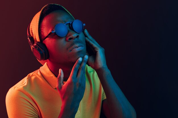 El joven guapo serio hipster triste escuchando música con auriculares en estudio negro con luces de neón.