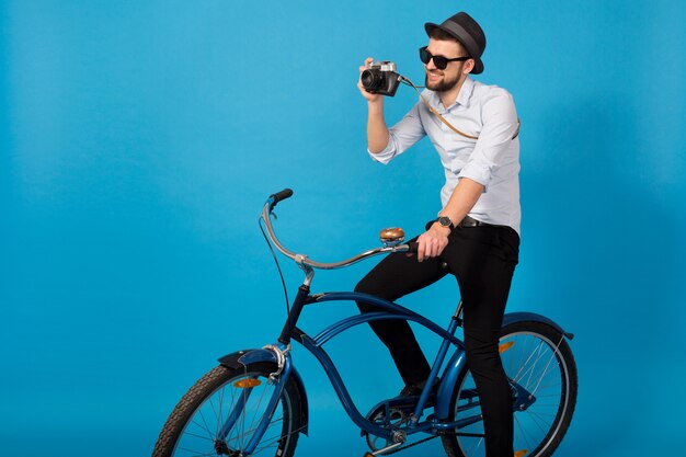 Joven guapo sonriente feliz viajando en bicicleta hipster, sosteniendo una cámara de fotos vintage sobre fondo azul de estudio, vistiendo camisa, sombrero y gafas de sol, fotógrafo tomando fotografías
