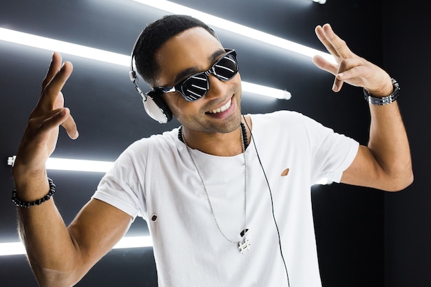 Joven guapo sonriente hipster negro en traje blanco escuchando música en auriculares y bailando al estilo hip hop en discoteca, divirtiéndose